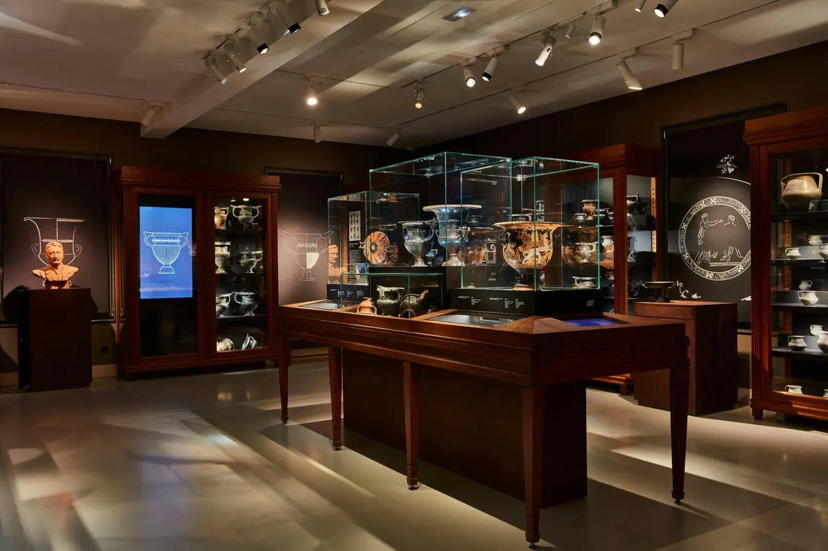 Une exposition dans un musée avec des vitrines en verre contenant des poteries et des objets anciens, éclairés par des projecteurs dans une pièce sombre.