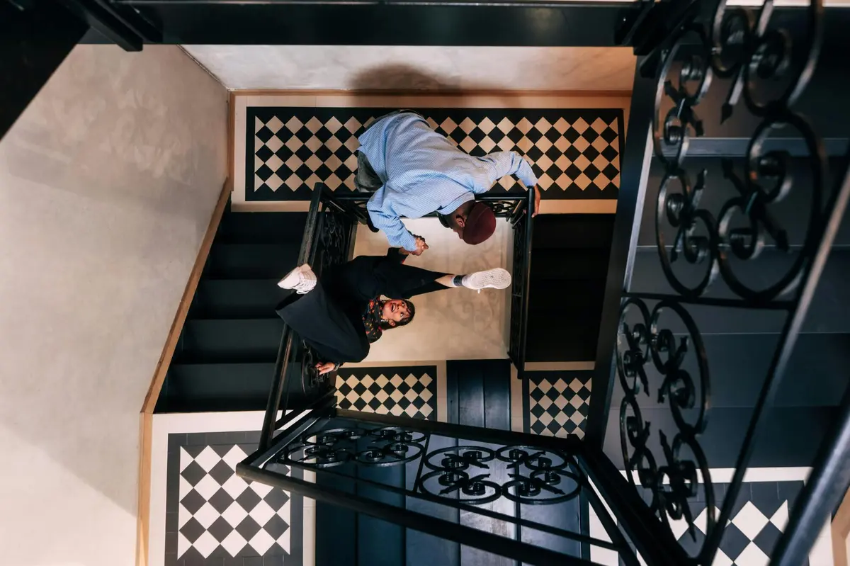 Deux personnes interagissent sur un escalier décoratif au sol carrelé noir et blanc, vu d'en haut.