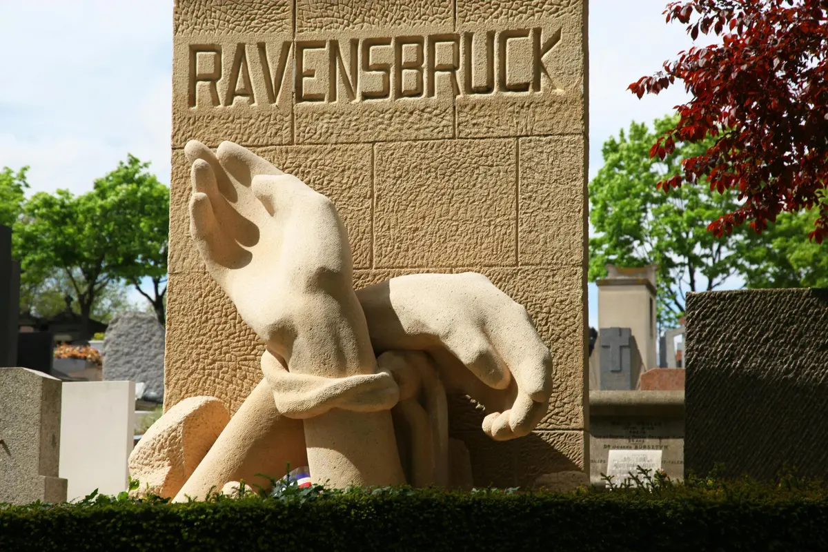 Un monument en pierre affiche le mot RAVENSBRUCK avec deux grandes mains sculptées, dont l'une est enchaînée, sur fond d'arbres et de pierres tombales.
