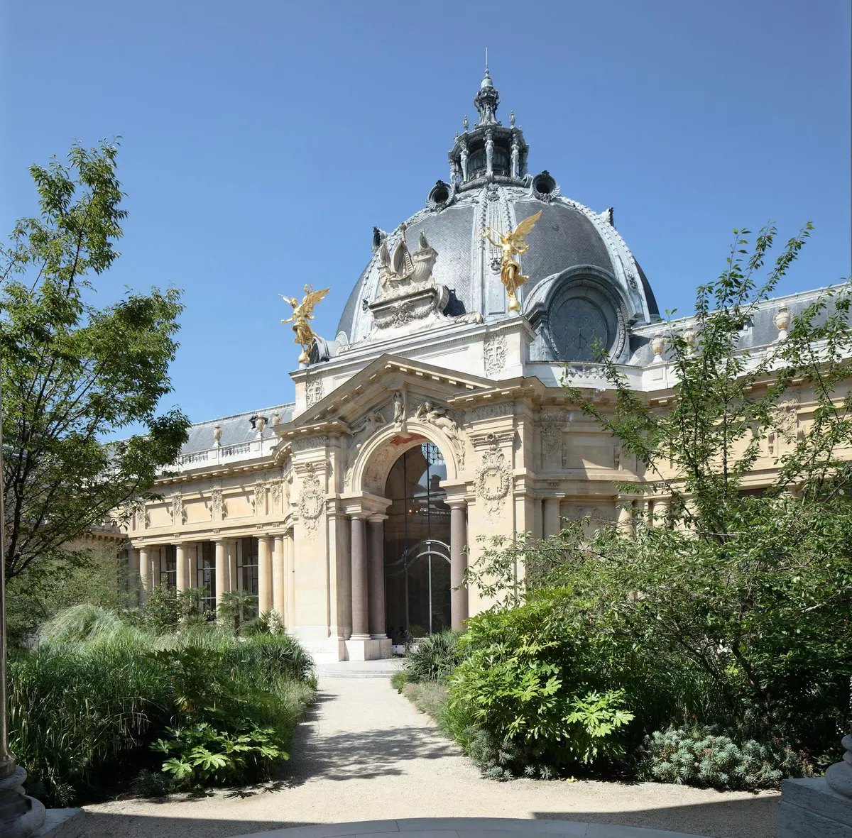 A historic building with a large dome, ornate architecture, and golden statues, surrounded by lush greenery under a clear blue sky.