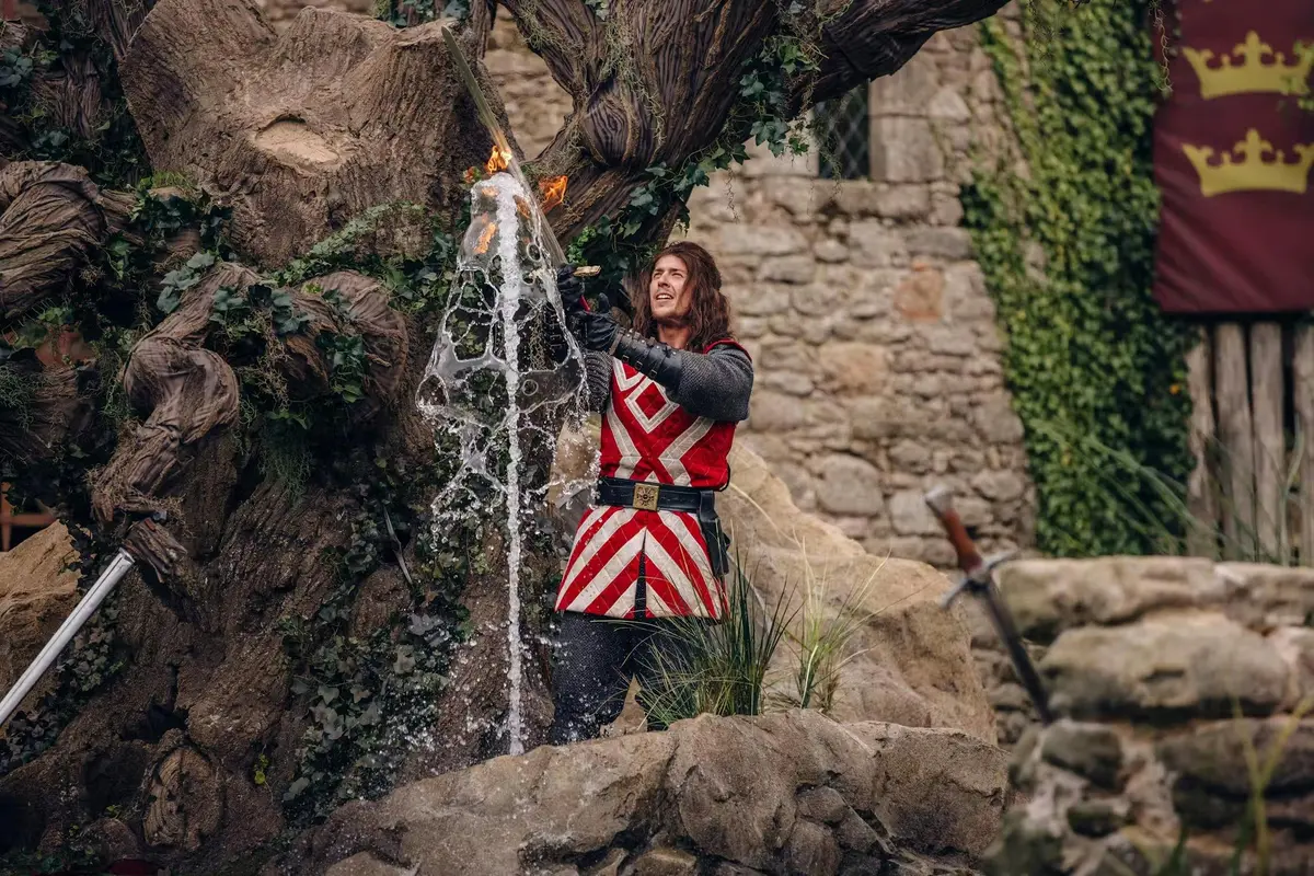 Une personne vêtue d'un costume médiéval sortant une épée enflammée d'une cascade d'eau, avec en arrière-plan un mur de pierre et de la végétation.