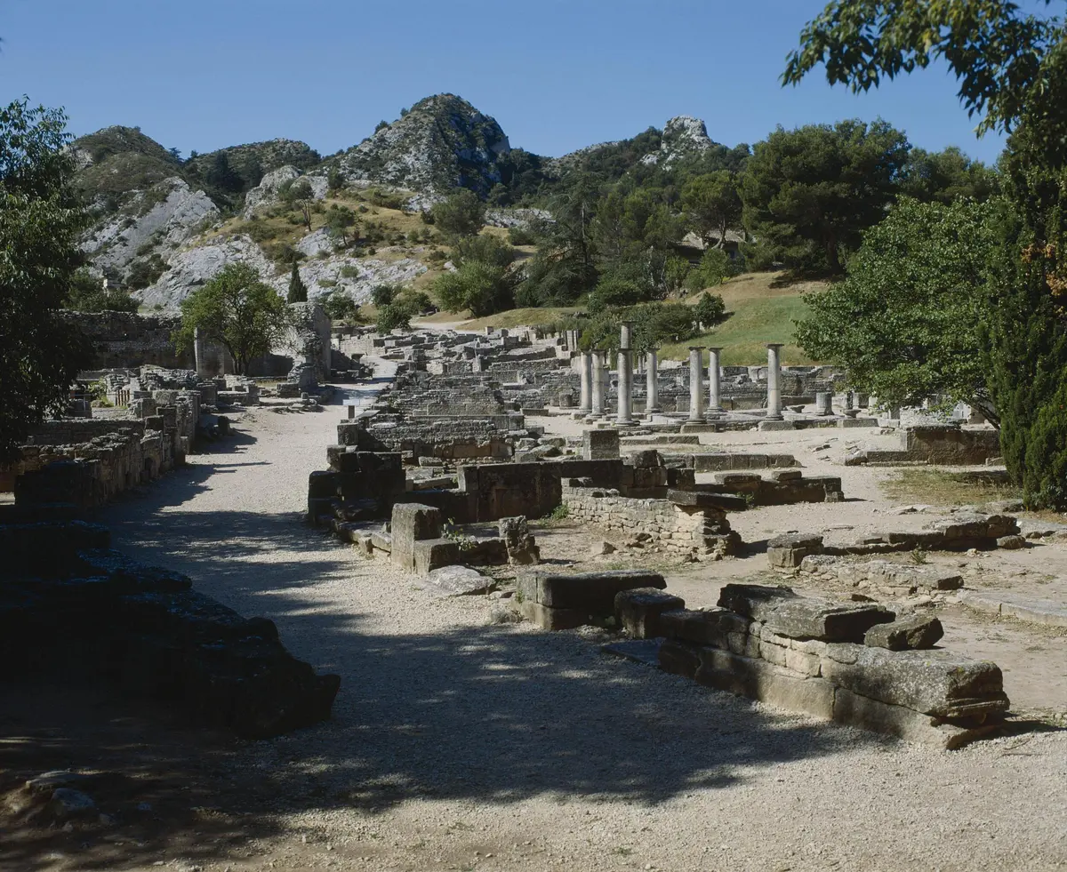 Des ruines anciennes, avec des structures en pierre brisée et des colonnes debout, sur fond de collines couvertes d'arbres.