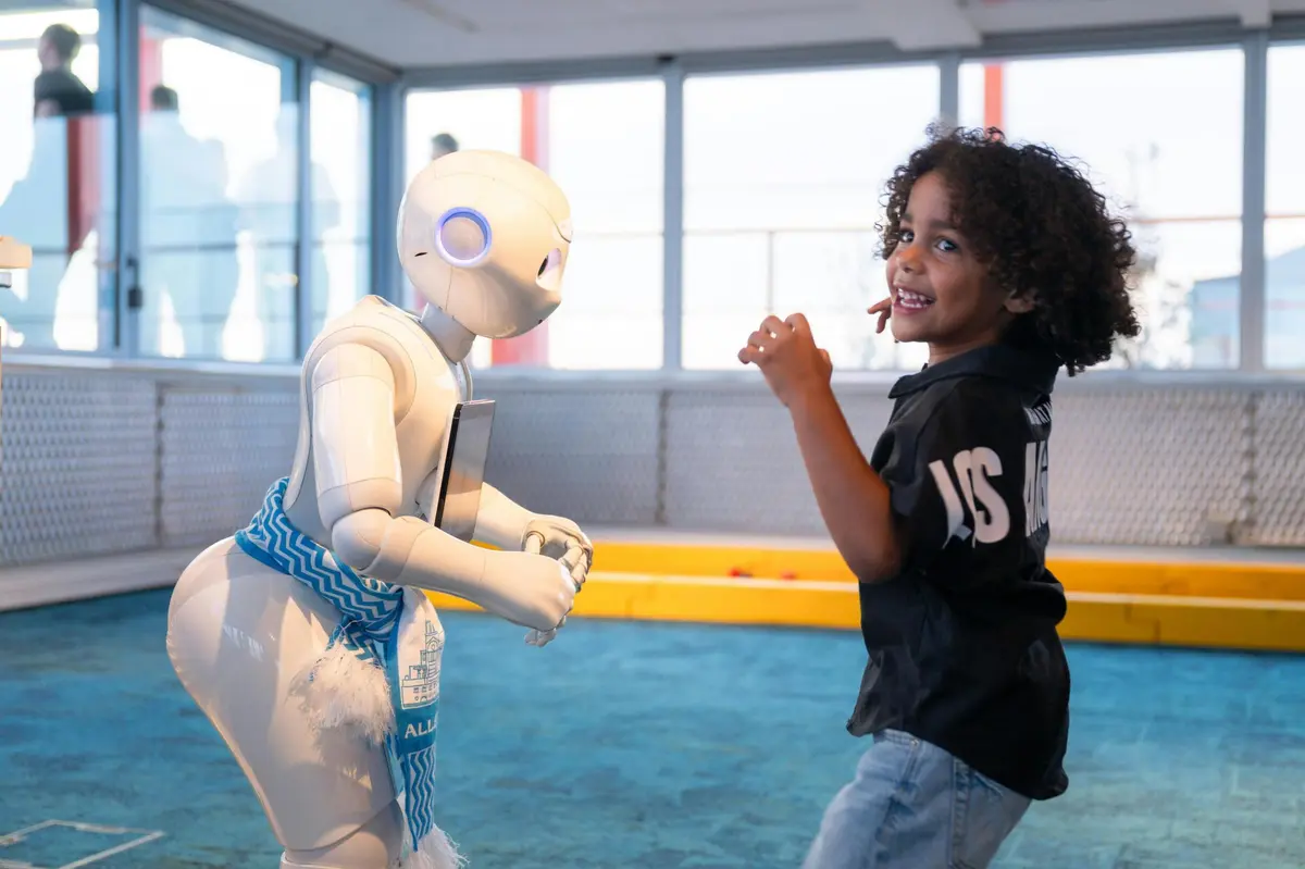 Un enfant aux cheveux bouclés, vêtu d'une chemise noire, sourit tout en interagissant avec un robot blanc dans une pièce dotée de grandes fenêtres.