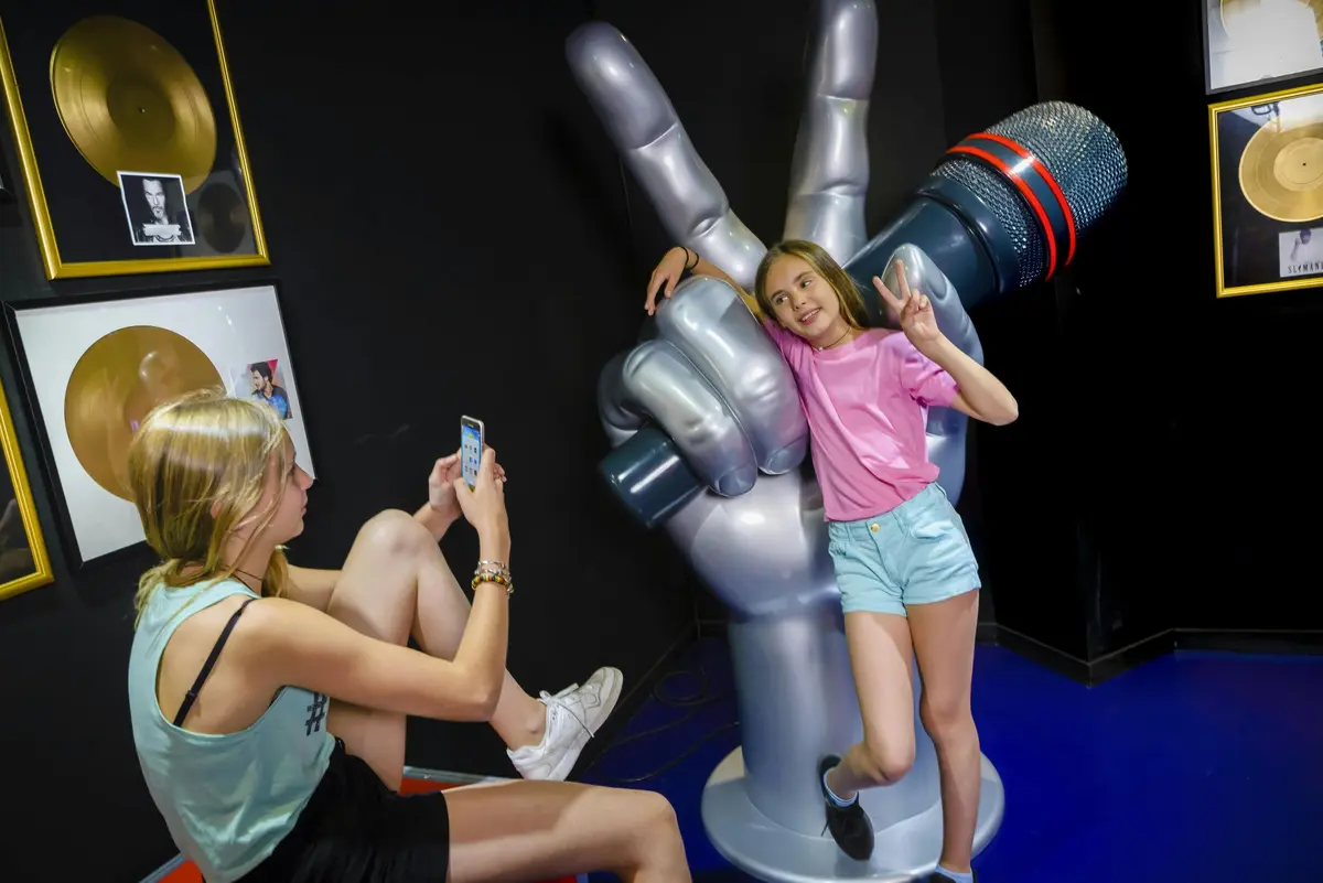 Une jeune fille pose avec un signe de paix près d'une grande sculpture représentant une main tenant un microphone, tandis qu'une autre personne la prend en photo.