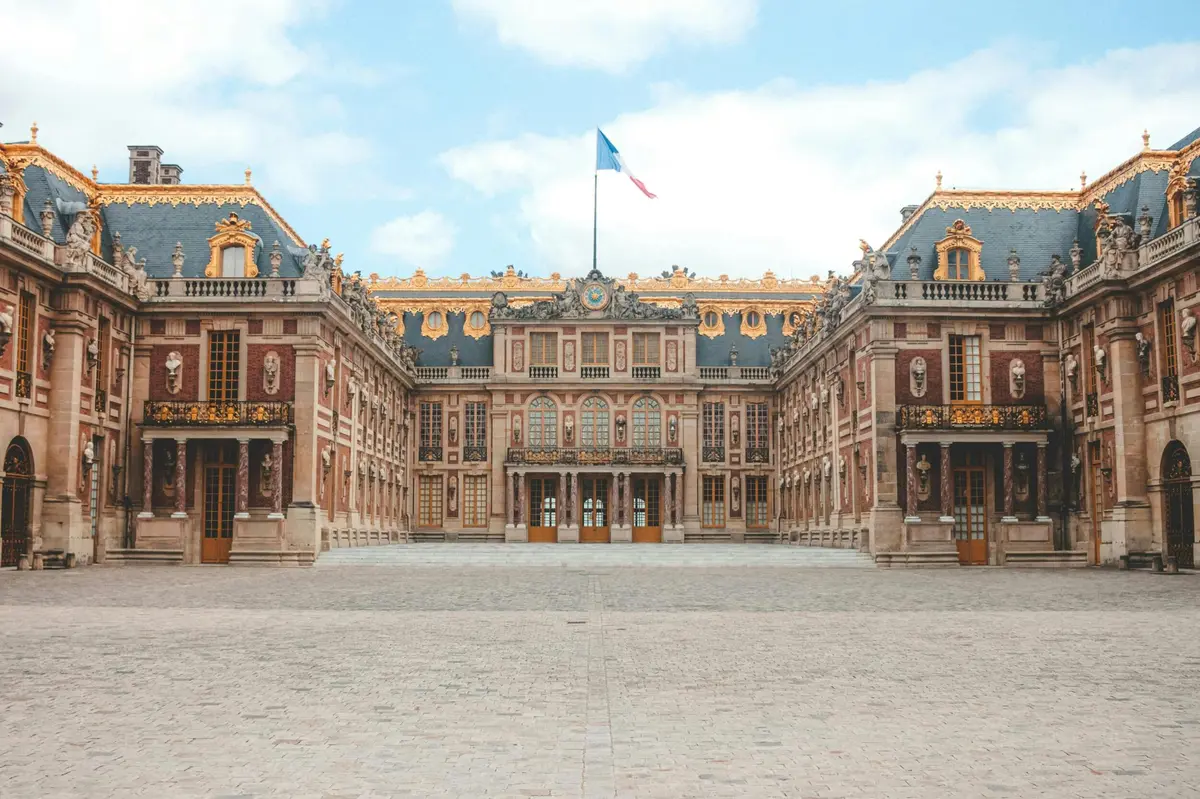 Un grand palais à l'architecture ornée, aux accents dorés et surmonté d'un drapeau français, vu d'une cour pavée.