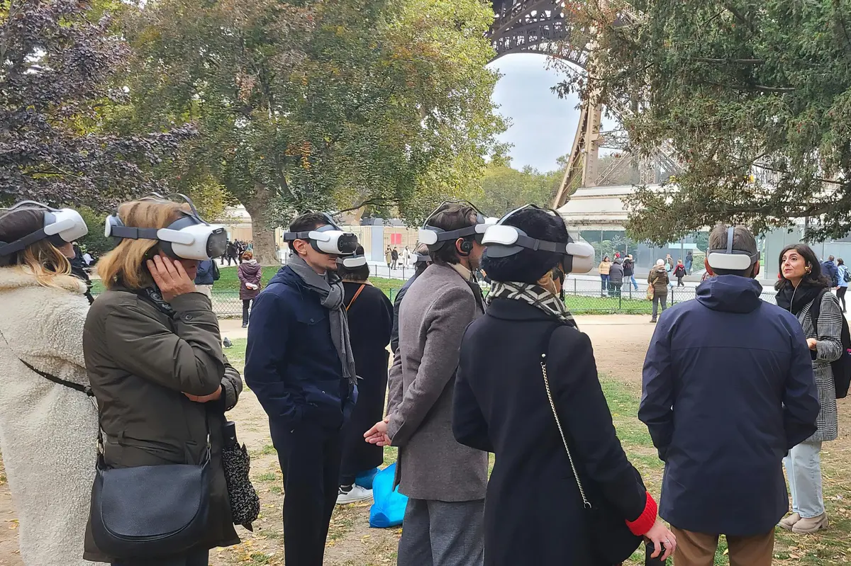 A group of people wearing VR headsets standing in a park near the Eiffel Tower, with a person addressing the group.