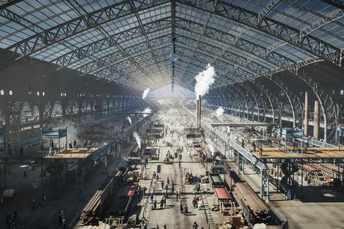 Un vaste hall industriel au toit voûté, grouillant de monde et de machines, d'où s'échappent de la fumée et de la vapeur.