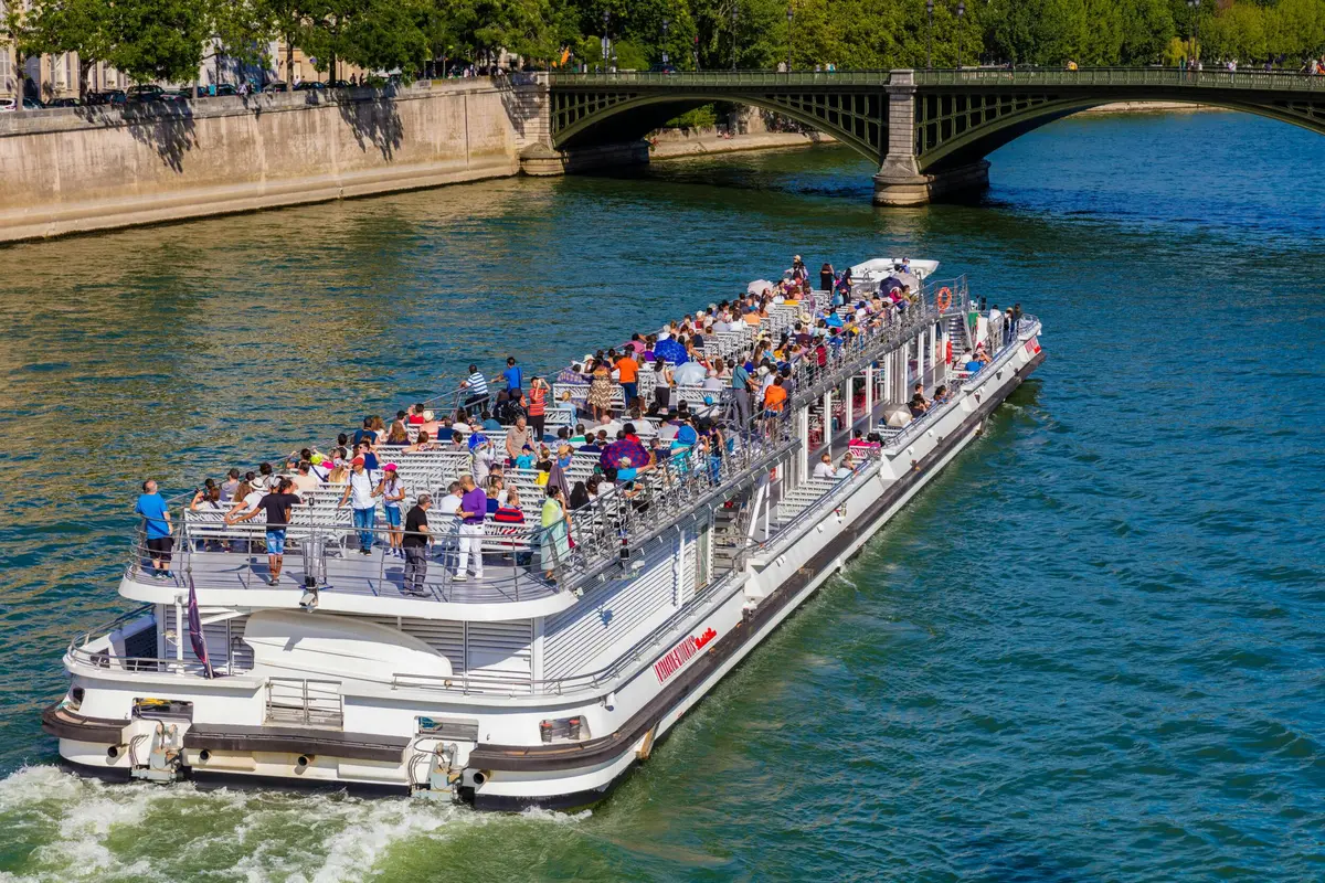 Un bateau de touristes bondé navigue sur une rivière près d'un pont en pierre avec de la verdure en arrière-plan.
