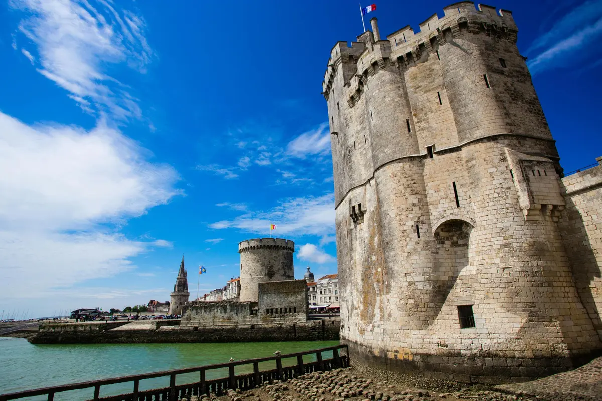 Deux tours médiévales au bord de l'eau par un ciel bleu clair. Des bâtiments et une flèche d'église sont visibles à l'arrière-plan.