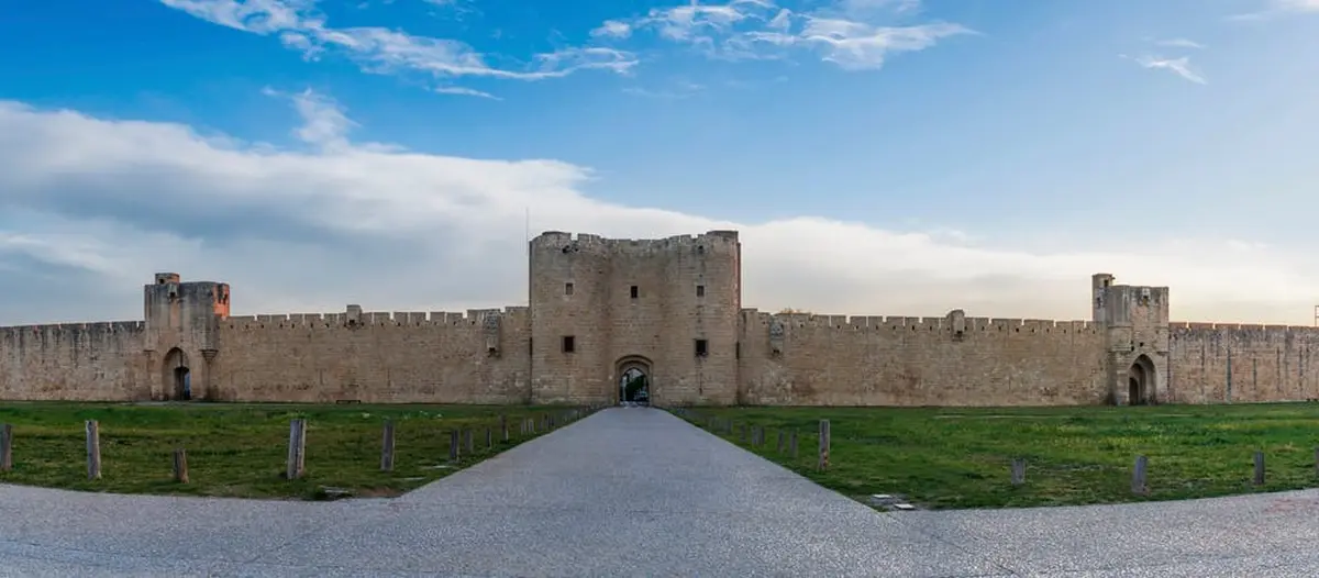 Forteresse en pierre avec une grande porte centrale, flanquée de hauts murs et de tours, sous un ciel bleu partiellement nuageux. Un large chemin mène à la porte.