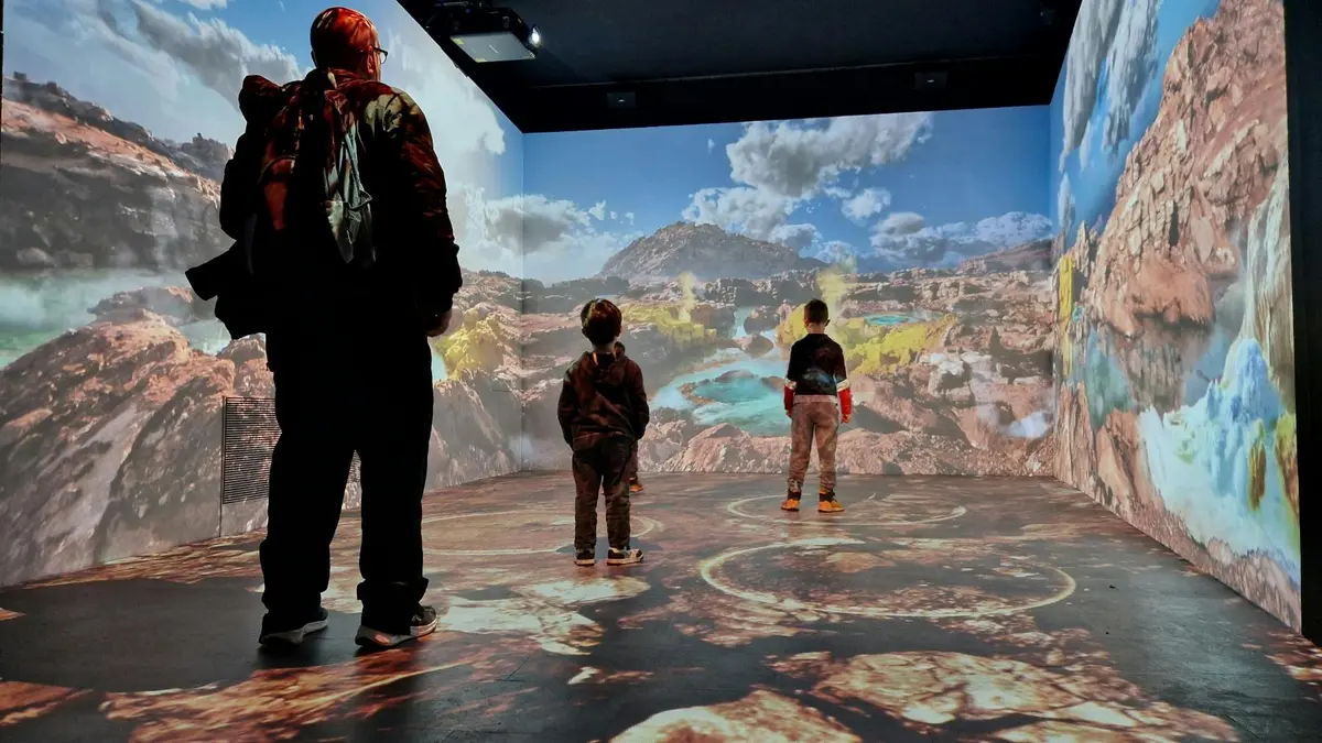 Trois personnes, dont deux enfants, se trouvent dans une pièce où un paysage désertique détaillé est projeté sur les murs et le sol.