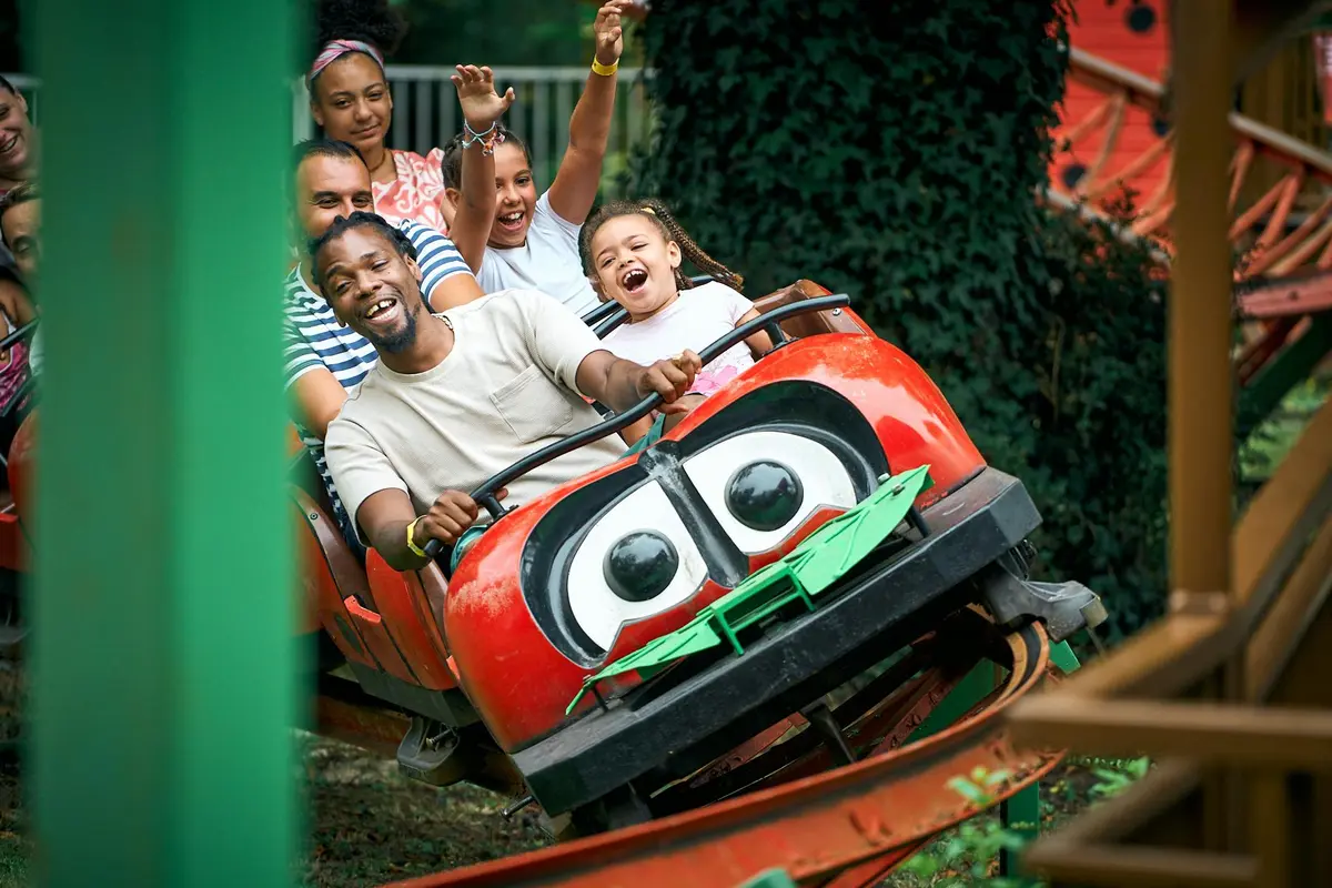 Groupe de personnes souriant et levant les mains tout en montant dans un wagon de montagnes russes rouge avec des yeux de dessins animés.