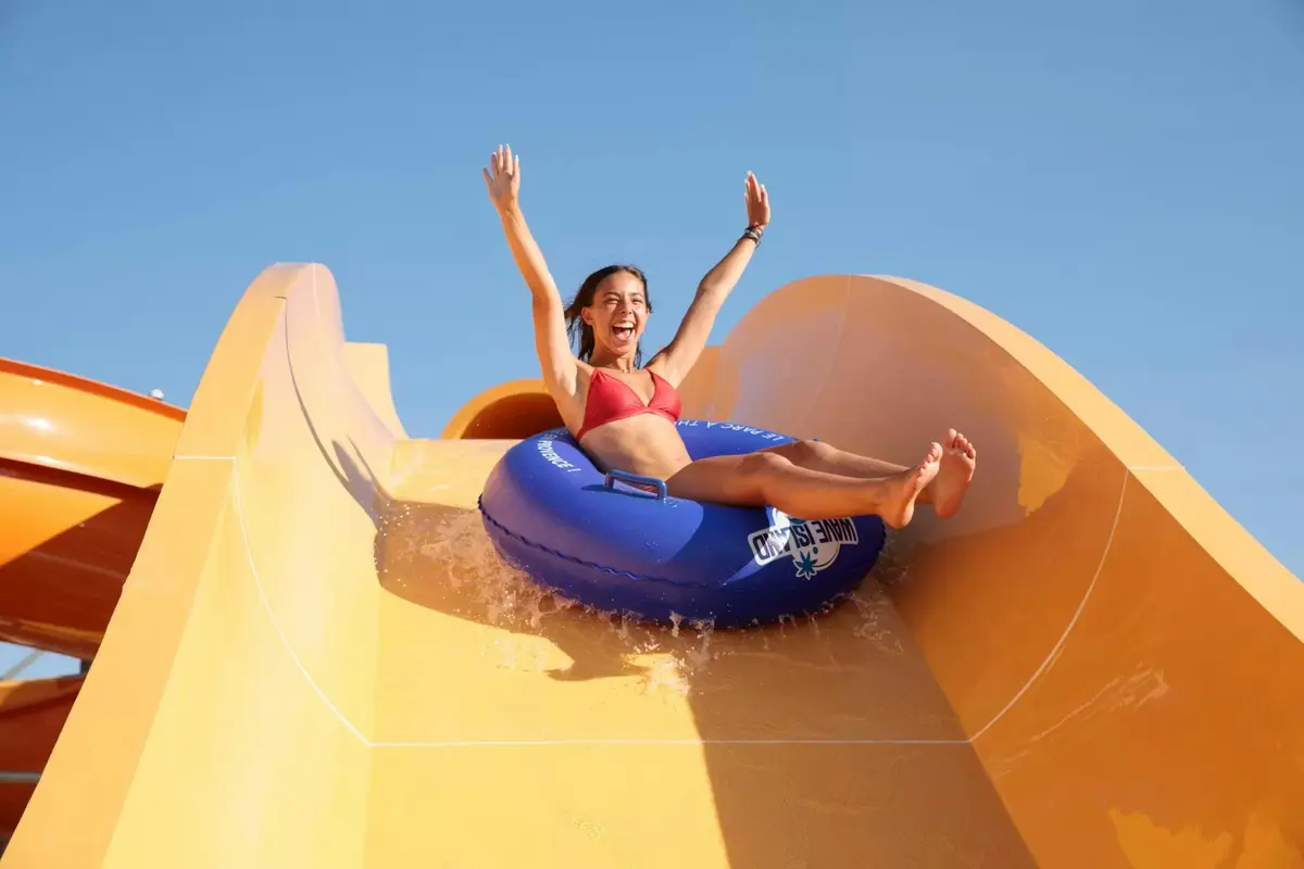 Une personne vêtue d'un maillot de bain rouge dévale un toboggan aquatique jaune sur une bouée bleue, en levant les bras avec joie.