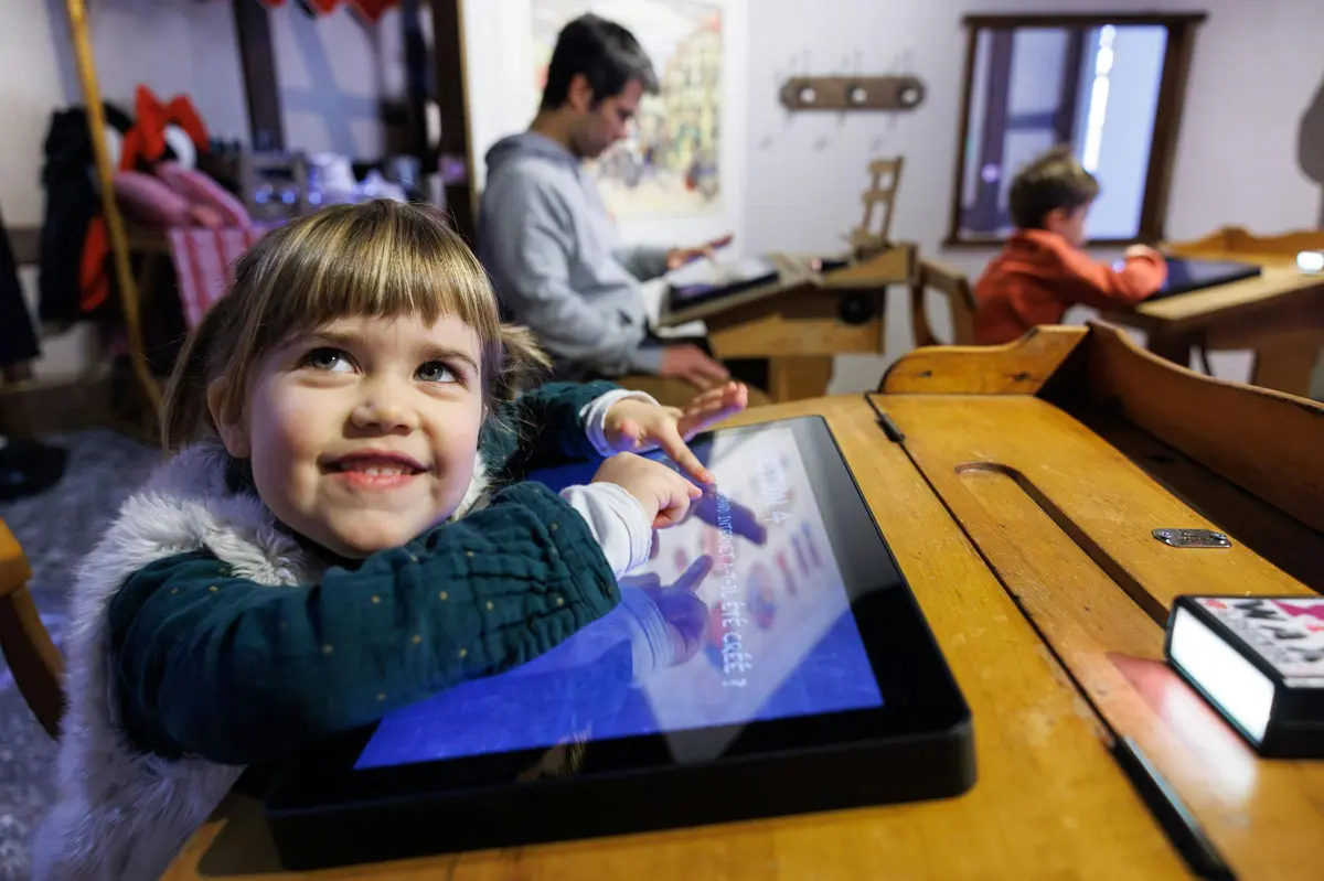 Une jeune fille qui sourit et montre du doigt un écran tactile, avec un adulte en train de lire à l'arrière-plan.