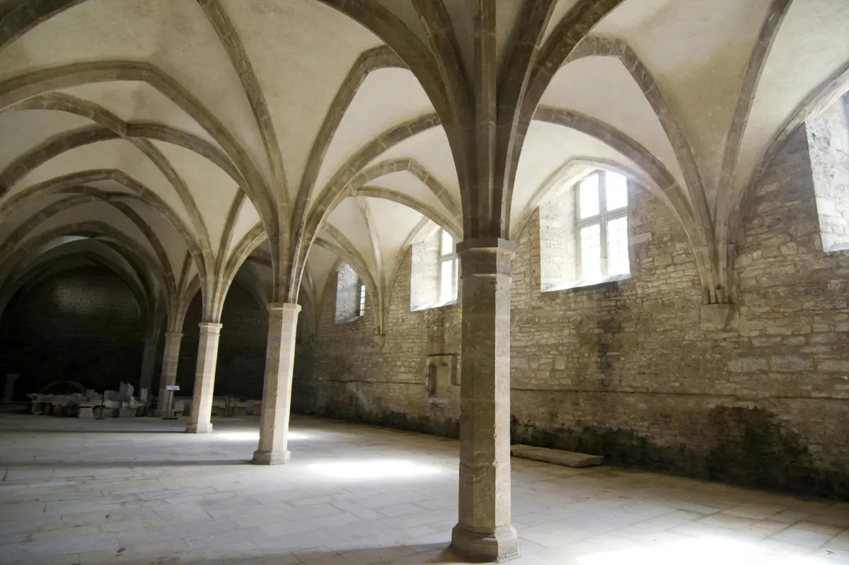 Hall en pierre avec de hauts plafonds voûtés, plusieurs colonnes et de grandes fenêtres. La lumière pénètre à flots et illumine l'espace.