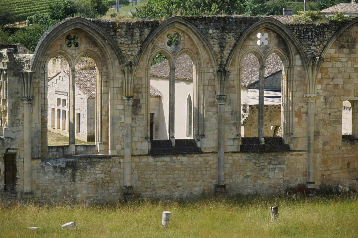 Ruines d'un bâtiment en pierre avec de grandes fenêtres cintrées et un premier plan herbeux. D'autres bâtiments sont visibles à travers les arches.