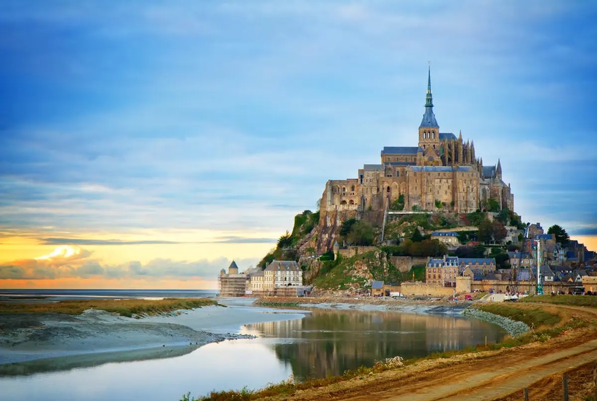 Abbaye médiévale et bâtiments sur une île rocheuse entourée d'eau, avec un pont reliant le continent au coucher du soleil.