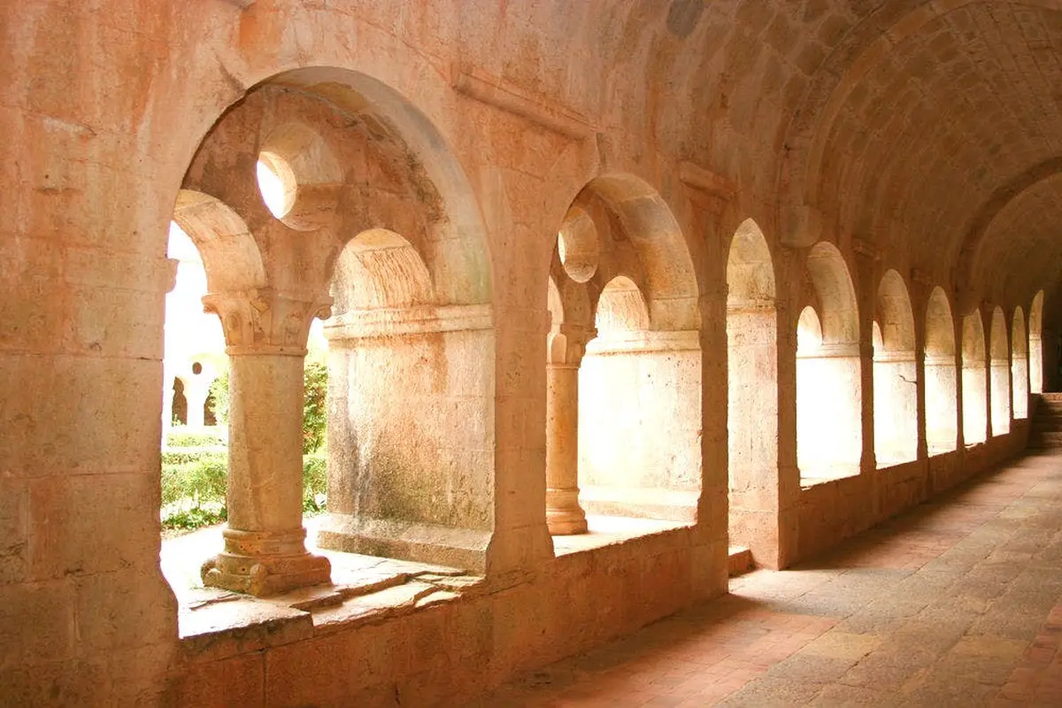 Allée en pierre avec des ouvertures en arc, des colonnes et une cour ensoleillée avec de la verdure en arrière-plan.