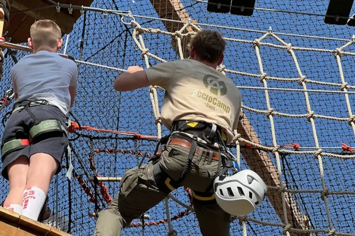 Personne dans un harnais grimpant à un filet de corde dans un parc d'aventure en plein air, avec un casque attaché à la ceinture.