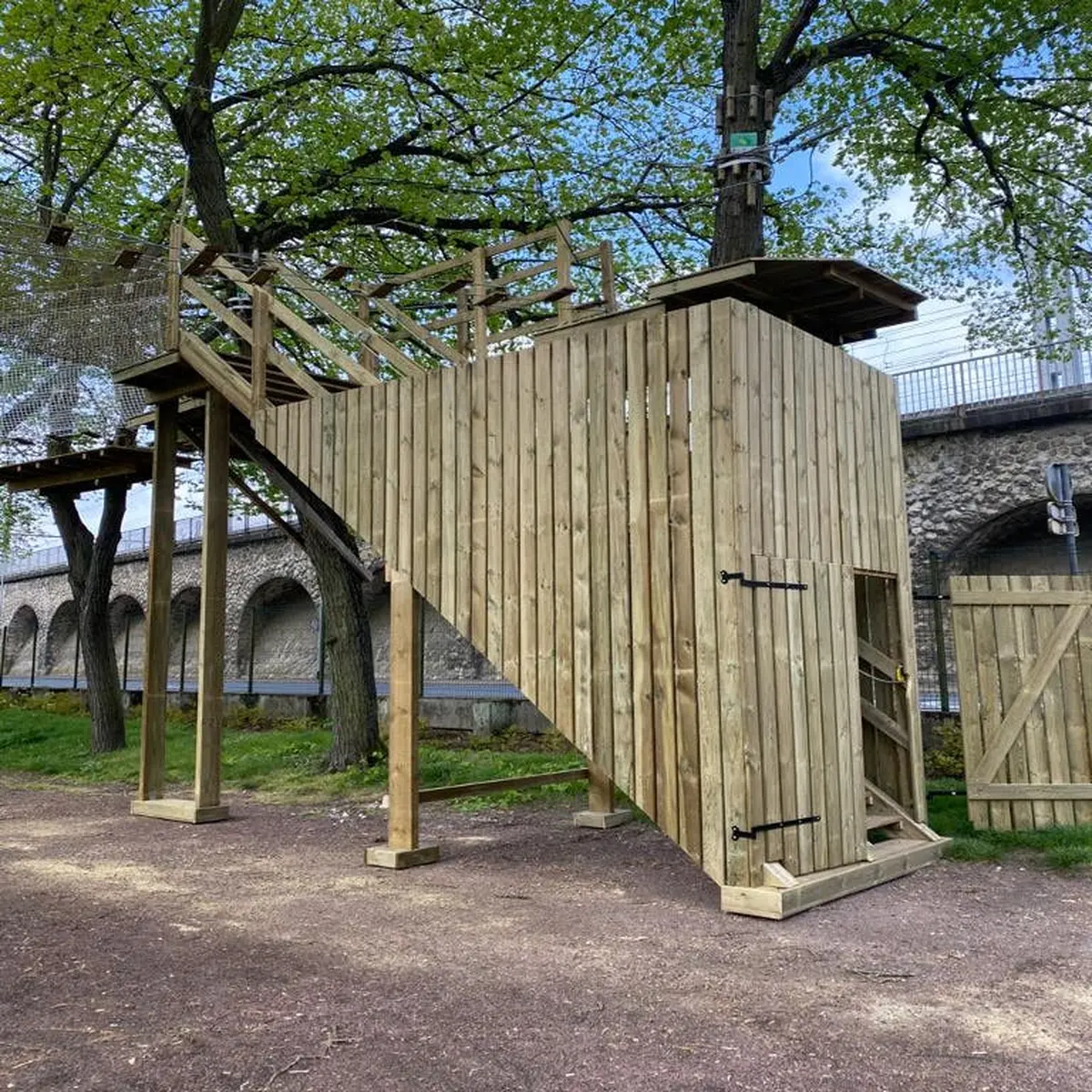 Structure de plate-forme en bois avec des escaliers et une porte, soutenue par des poteaux. Située à l'extérieur, près d'arbres et d'un mur de pierre avec des arches.
