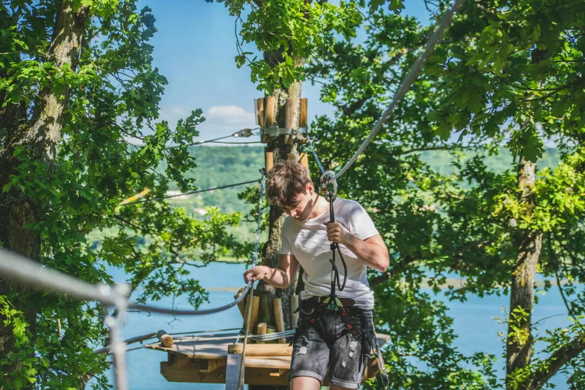 Une personne portant un harnais navigue sur un parcours de cordes dans une forêt luxuriante et feuillue, avec un lac et des collines en arrière-plan.