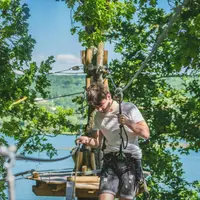 Une personne portant un harnais navigue sur un parcours de cordes dans une forêt luxuriante et feuillue, avec un lac et des collines en arrière-plan. &copy; AccroCamp