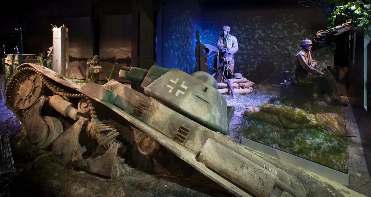 Exposition muséale présentant un char d'assaut militaire endommagé et des mannequins habillés comme des soldats dans une scène de tranchées de la Seconde Guerre mondiale.