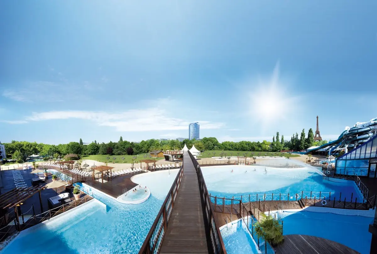 Grand parc aquatique en plein air avec des piscines, une passerelle en bois et la verdure environnante. Immeuble de grande hauteur en arrière-plan sous un ciel lumineux.