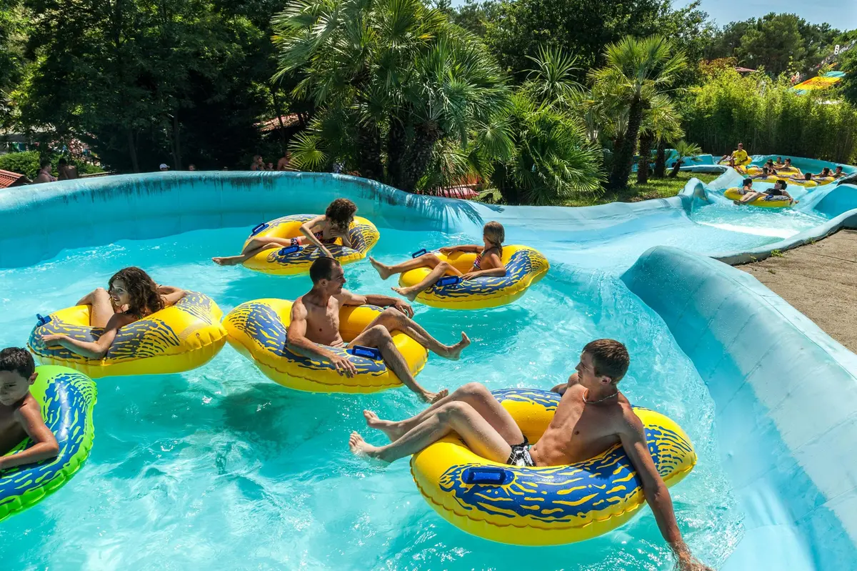 Des personnes flottant sur des bouées jaunes dans la piscine d'un parc aquatique, au milieu d'une végétation luxuriante et de palmiers.