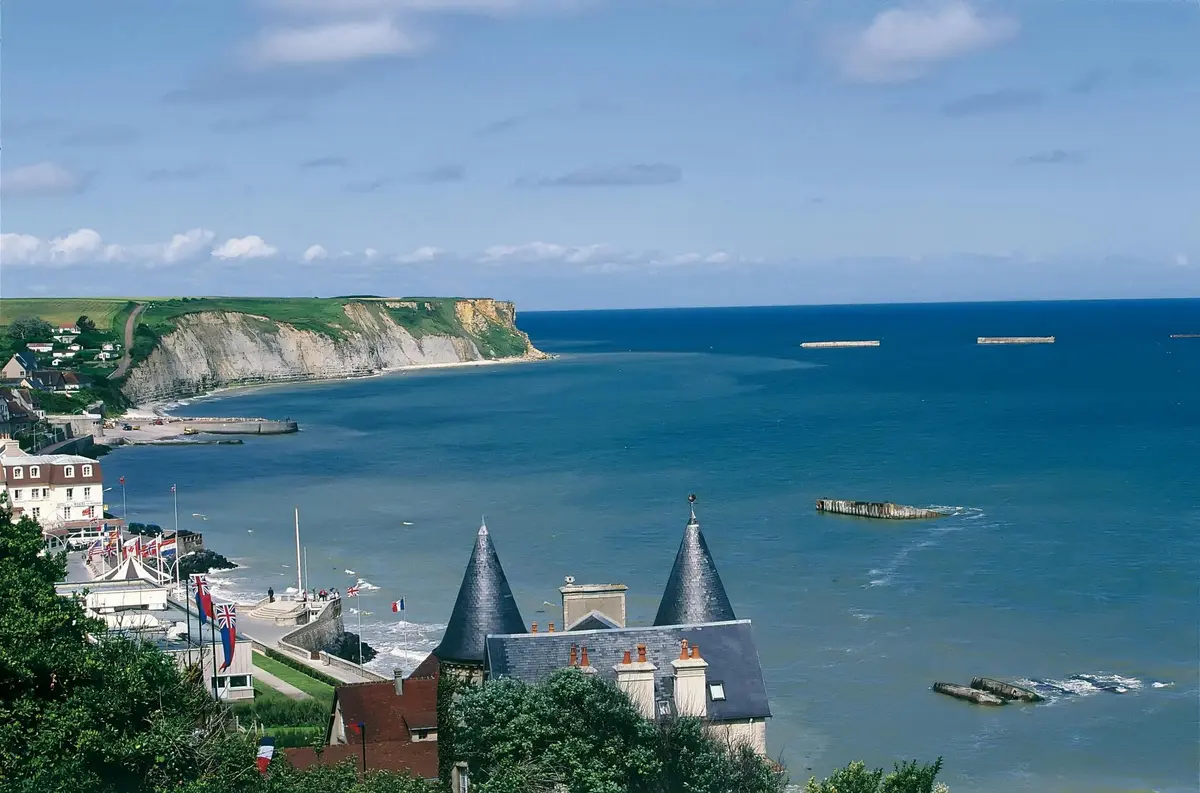 Scène côtière avec l'océan bleu, des falaises rocheuses au loin et un bâtiment à toit d'ardoise avec des tours pointues au premier plan.