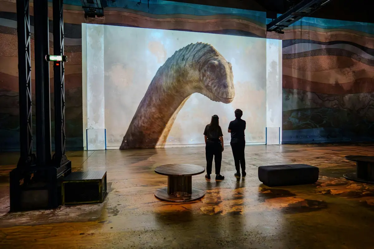 Deux personnes se tiennent devant un grand écran affichant l'image de la tête et du cou d'un dinosaure dans une pièce sombre et spacieuse.