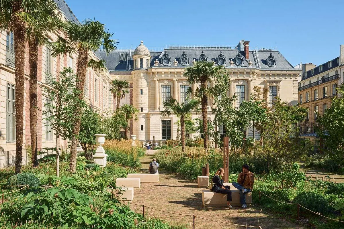 Un jardin agrémenté de plantes et de palmiers devant un bâtiment de style classique. Des gens sont assis sur des bancs et se promènent.