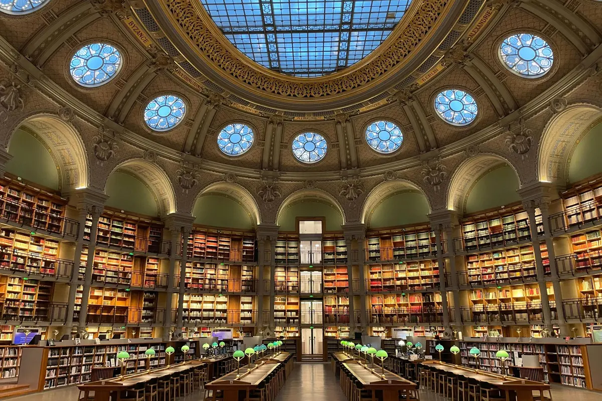 Une grande bibliothèque avec un plafond en verre bombé, des étagères circulaires et des rangées de tables avec des lampes de lecture vertes.