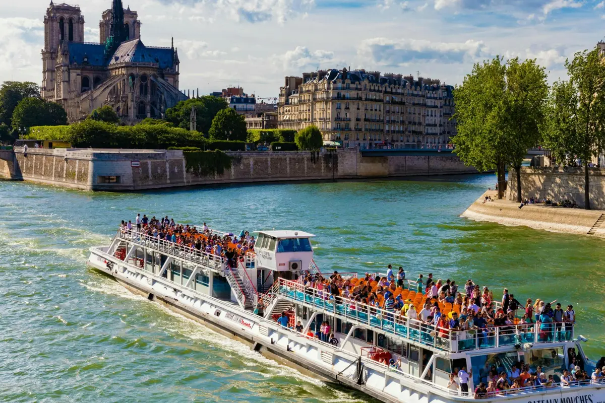 Un bateau d'excursion bondé navigue sur une rivière près de bâtiments historiques et d'arbres sous un ciel partiellement nuageux.