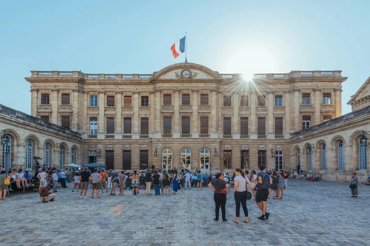 Un bâtiment historique surmonté d'un drapeau français, un soleil radieux et une foule de personnes dans la cour.