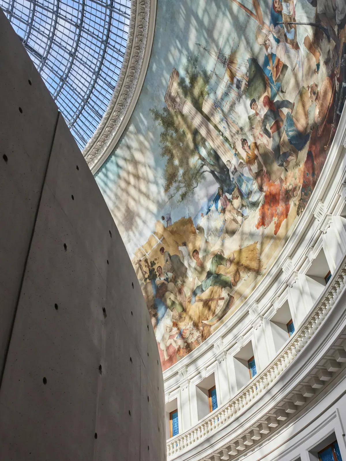 Un mur incurvé à l'intérieur d'un bâtiment avec une grande peinture murale détaillée représentant des personnes et la nature sous un plafond de verre en forme de dôme.
