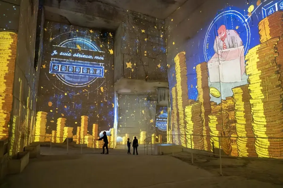 Une installation artistique immersive dans un espace caverneux, avec des projections de pièces d'or, d'étoiles et d'un homme à un bureau.