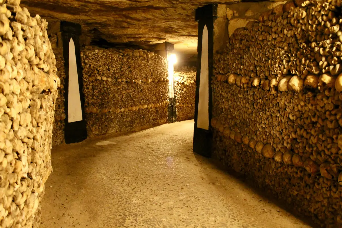 Un passage souterrain faiblement éclairé dont les murs sont faits d'os et de crânes soigneusement empilés, avec des piliers noirs décoratifs.