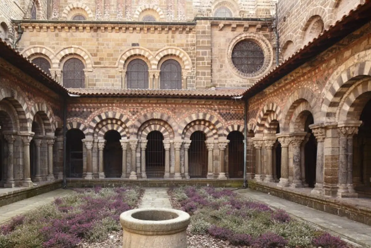 Cour médiévale avec des arcs et des colonnes en pierre, avec un puits central en pierre et des plantes à fleurs au premier plan.