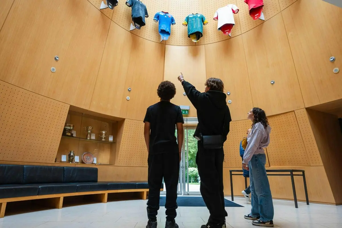 Trois personnes se tiennent dans une pièce circulaire aux murs en bois, observant les maillots accrochés en hauteur sur le mur et les trophées posés sur des étagères.