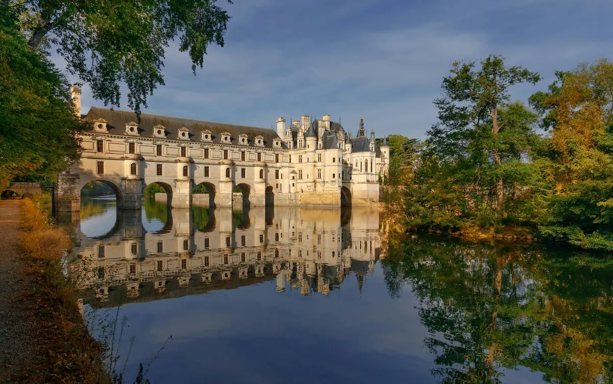 Un château aux ponts arqués enjambe une rivière calme, avec des reflets clairs de la structure et des arbres environnants dans l'eau.