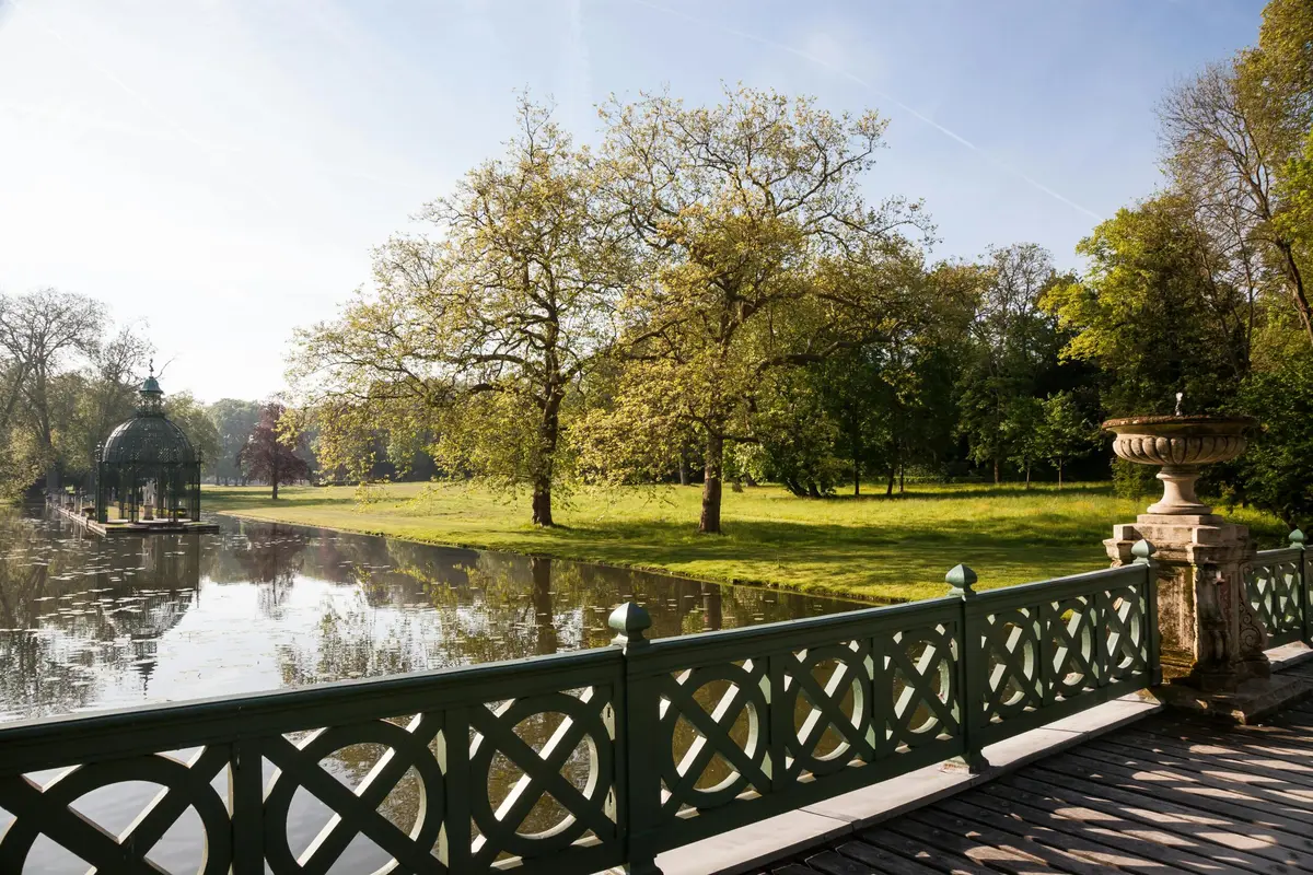 Parc verdoyant avec une balustrade décorative au premier plan, de grands arbres et leur reflet dans un étang calme sous un ciel clair.