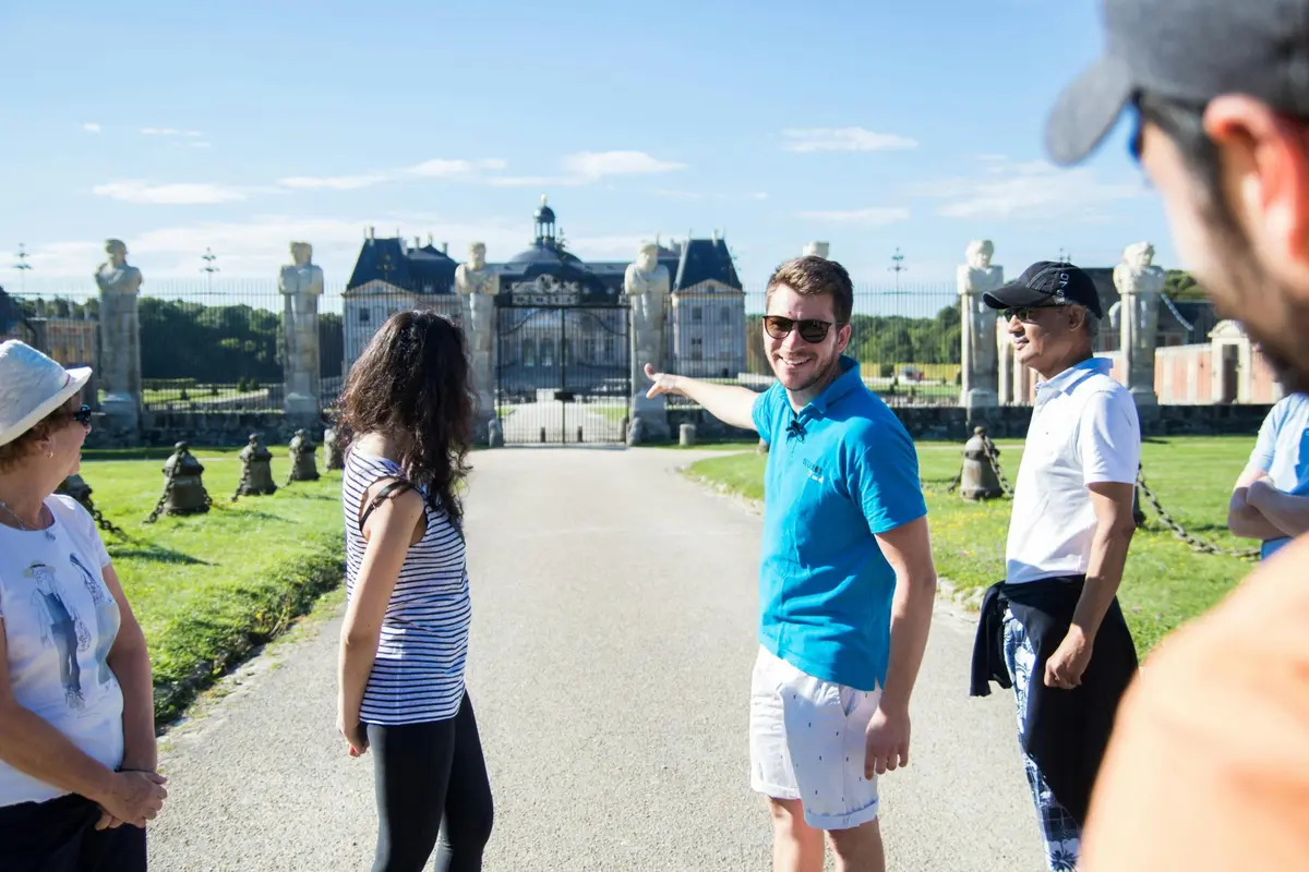 Un groupe de personnes à l'extérieur, dont l'une montre du doigt un bâtiment situé à l'arrière-plan, par une journée ensoleillée.