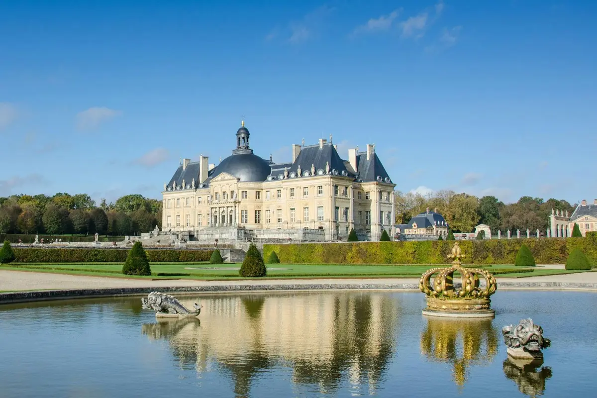 A grand palace with blue-domed roofs reflected in a large ornamental pond, featuring sculptures and a crown-shaped fountain.