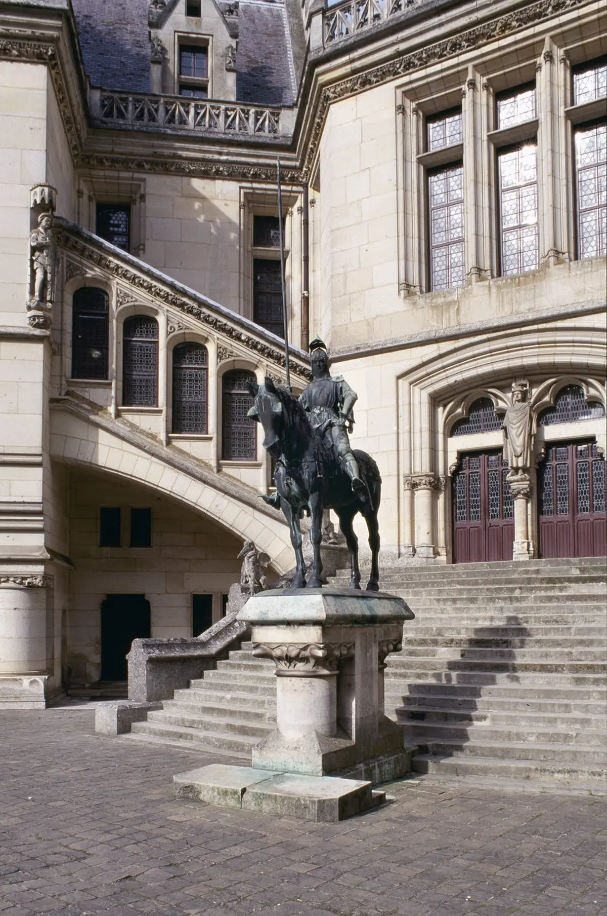 La statue en pierre d'un chevalier à cheval est placée devant un bâtiment orné d'escaliers, d'arcs et de grandes fenêtres.