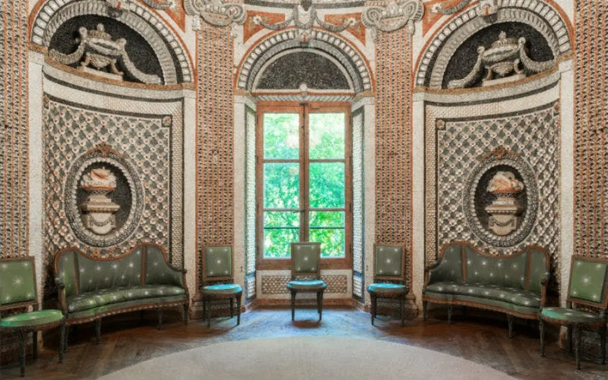 Une pièce ornée de motifs muraux complexes, de quatre chaises vertes et d'une grande fenêtre donnant sur la verdure luxuriante à l'extérieur.