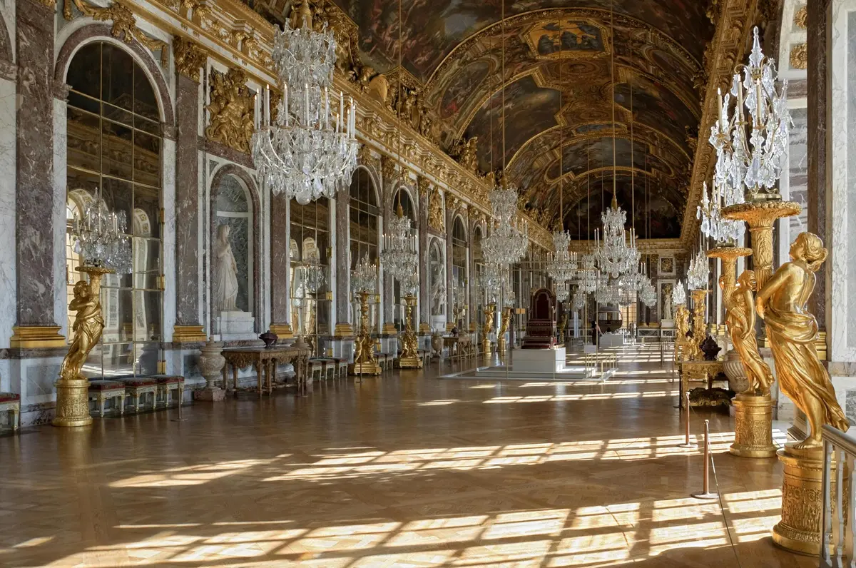 Ornate hall with high arched ceilings, large chandeliers, marble statues, and tall windows letting in sunlight.