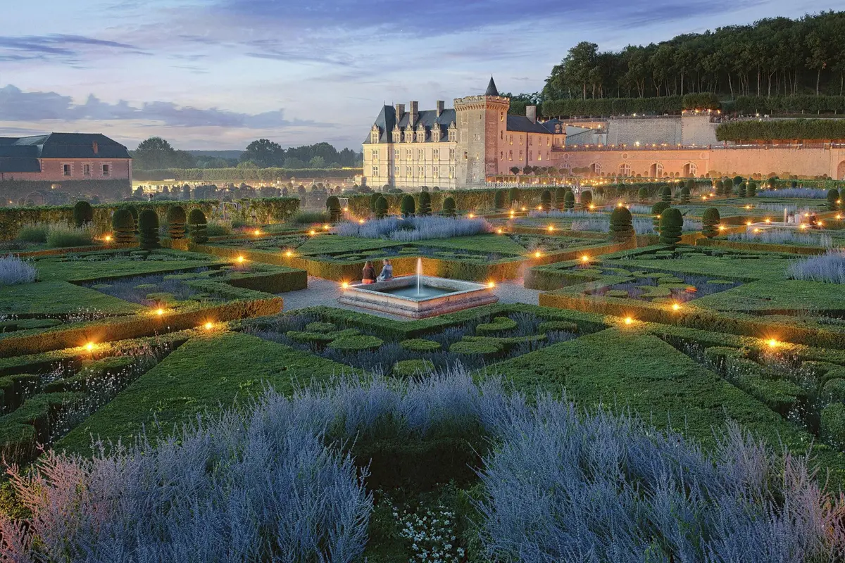 Jardin formel avec des haies géométriques et des allées éclairées devant un château historique au crépuscule. Deux personnes sont assises près d'une fontaine centrale.