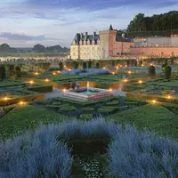 Jardin formel avec des haies géométriques et des allées éclairées devant un château historique au crépuscule. Deux personnes sont assises près d'une fontaine centrale. DR