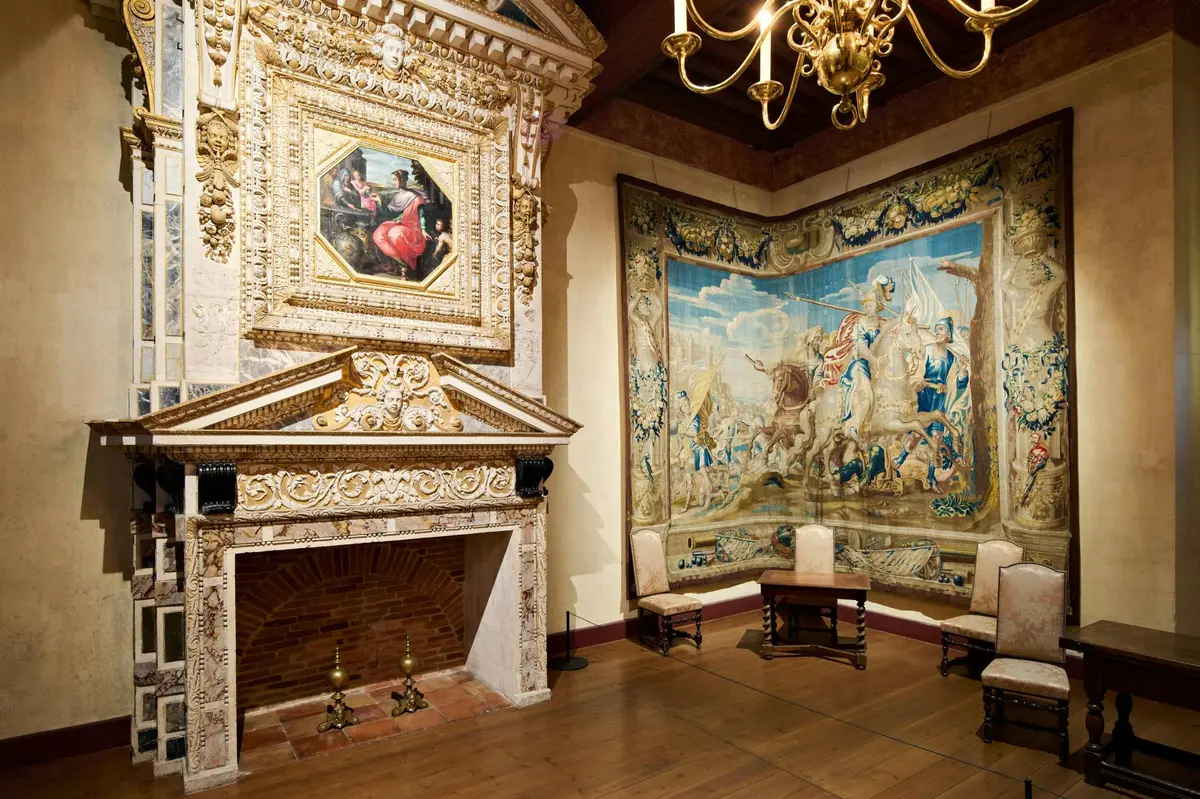 Salle élégante avec cheminée ornée, tableau encadré, grande tapisserie représentant une scène historique, chaises en bois et lustre.